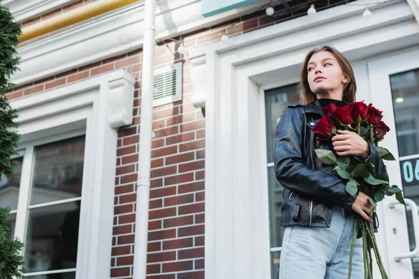 Mulher jovem e na moda em jaqueta de couro segurando rosas vermelhas na rua urbana na Europa — Fotografia de Stock