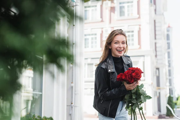 Mujer joven positiva en chaqueta de cuero con rosas rojas en la calle urbana en Europa - foto de stock