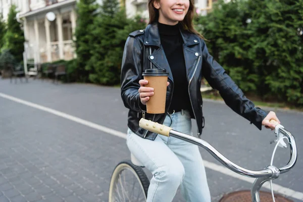 Vista recortada de la mujer feliz sosteniendo taza de papel mientras monta bicicleta en la calle urbana de Europa - foto de stock