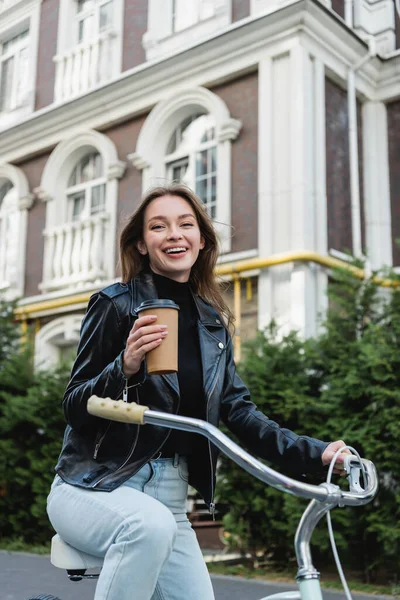 Mulher alegre segurando copo de papel ao andar de bicicleta na rua urbana da Europa — Fotografia de Stock