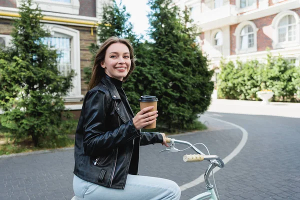 Young happy woman in leather jacket riding bicycle while holding paper cup — Stock Photo