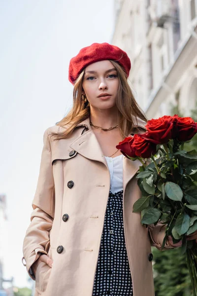 Élégante femme en trench coat et béret rouge tenant des roses et posant la main dans la poche à l'extérieur — Photo de stock