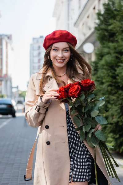 Щаслива молода жінка в траншеї пальто і червоний берет тримає букет троянд на міській вулиці Європи — стокове фото