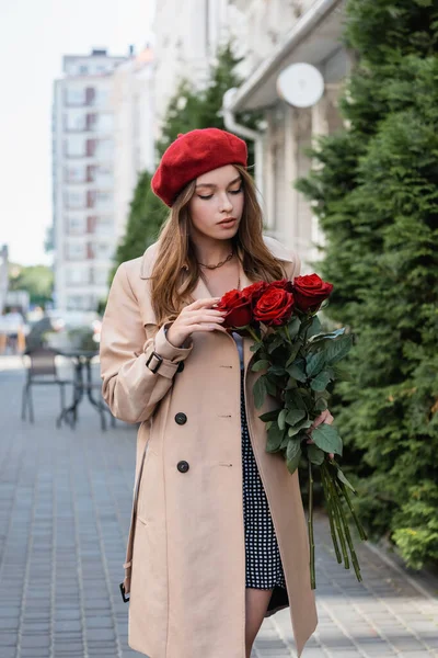 Bonita mujer en gabardina y boina roja mirando ramo de rosas en la calle urbana de Europa - foto de stock