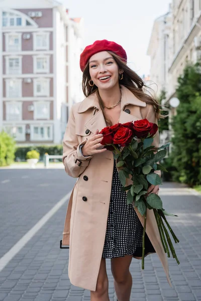 Cheerful young woman in trench coat and red beret holding bouquet of roses on urban street of europe — Stock Photo
