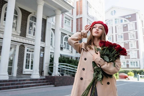 Mujer joven en gabardina sosteniendo rosas rojas y ajustando la boina en la calle urbana de Europa - foto de stock