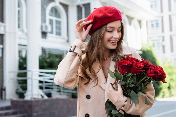 Happy young woman in trench coat holding red roses and adjusting beret on urban street of europe — Stock Photo