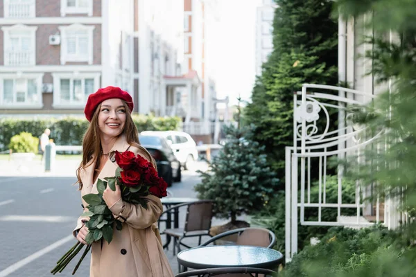 Mulher alegre em casaco de trincheira e boina vermelha segurando buquê de rosas na rua urbana da Europa — Fotografia de Stock