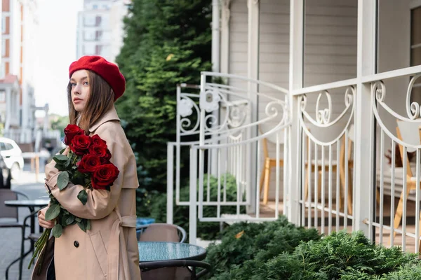 Joven bonita mujer en gabardina y boina sosteniendo rosas rojas en la calle urbana de Europa - foto de stock