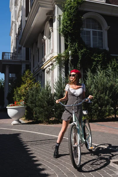 Full length of pretty woman sunglasses, red beret and dress standing near bicycle on urban street in europe — Stock Photo