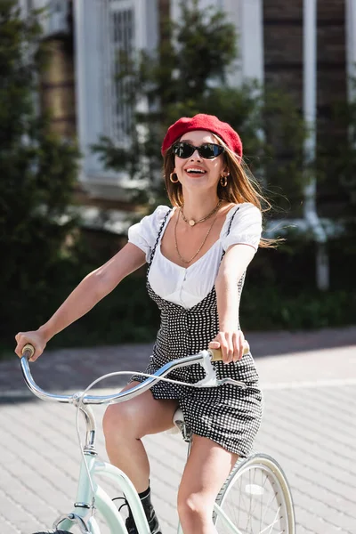 Felice giovane donna berretto rosso, occhiali da sole e vestito in bicicletta vicino alla costruzione in Europa — Foto stock