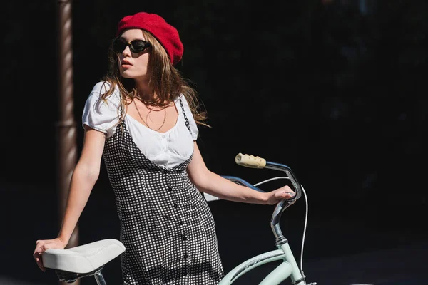 Красивая молодая женщина в солнечных очках, берете и платье стоя с велосипедом — стоковое фото