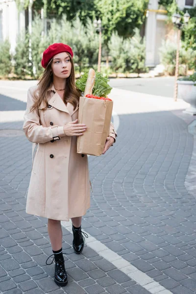 Повна довжина молодої жінки в бежевому траншеї пальто і червоний берет тримає паперовий мішок з продуктами на міській вулиці — стокове фото
