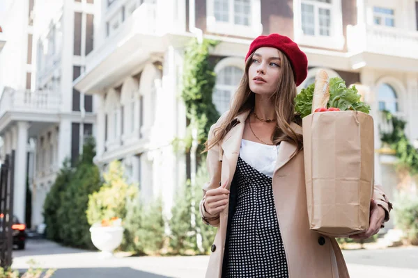 Junge Frau in beigem Trenchcoat und roter Baskenmütze hält Papiertüte mit Lebensmitteln auf der Straße — Stockfoto