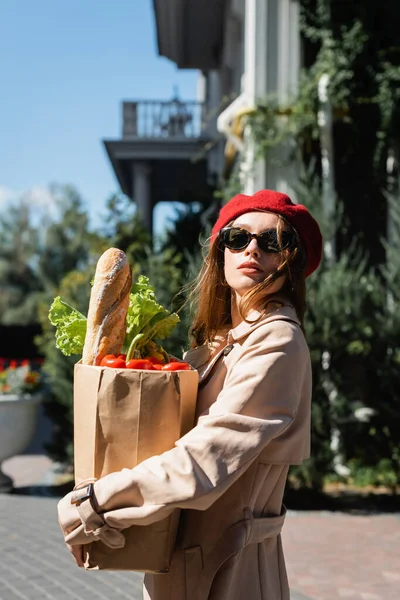 Junge Frau in beigem Trenchcoat, Sonnenbrille und roter Baskenmütze mit Papiertüte und Lebensmitteln — Stockfoto