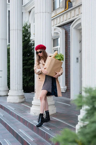 Piena lunghezza di giovane donna in trench beige, occhiali da sole e berretto rosso con sacchetto di carta con generi alimentari vicino all'edificio — Foto stock