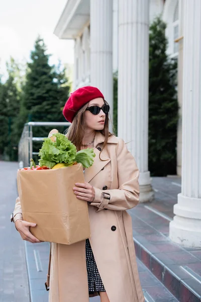 Junge Frau in Trenchcoat, Sonnenbrille und roter Baskenmütze hält Papiertüte mit Lebensmitteln in der Nähe des Gebäudes — Stockfoto