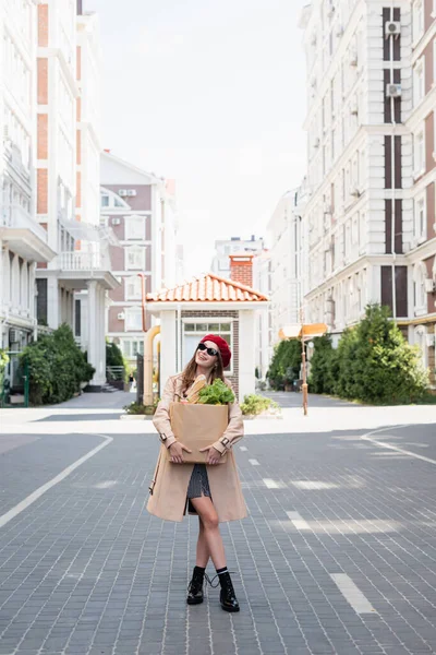 Full length of cheerful woman in trench coat, sunglasses and red beret holding paper bag with groceries on urban street — Stock Photo