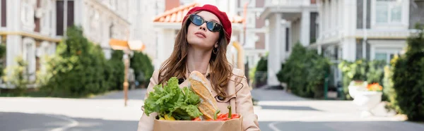 Femme rêveuse en trench coat beige, lunettes de soleil et béret tenant sac en papier avec épicerie sur la rue d'Europe, bannière — Photo de stock