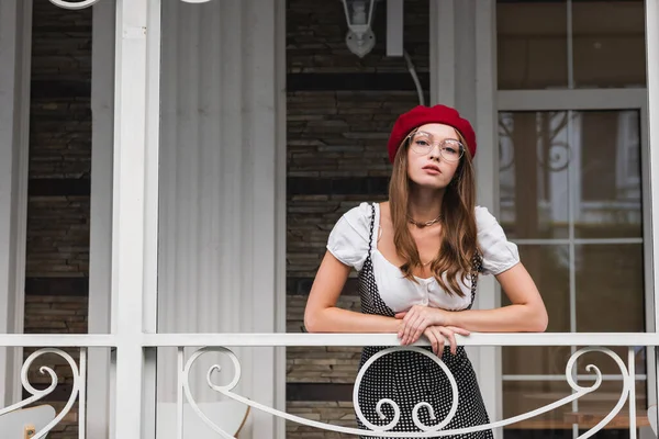 Trendy young woman in red beret and eyeglasses standing on balcony near house — Stock Photo