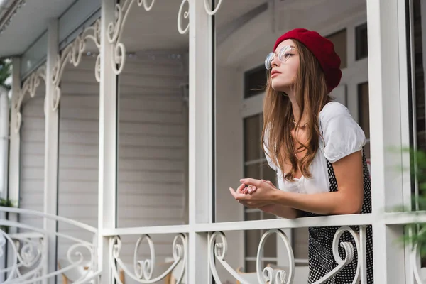 Young woman in red beret and eyeglasses standing on balcony and looking away — Stock Photo