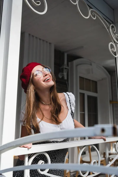 Jovem alegre em boina vermelha e óculos de pé na varanda perto da casa — Fotografia de Stock