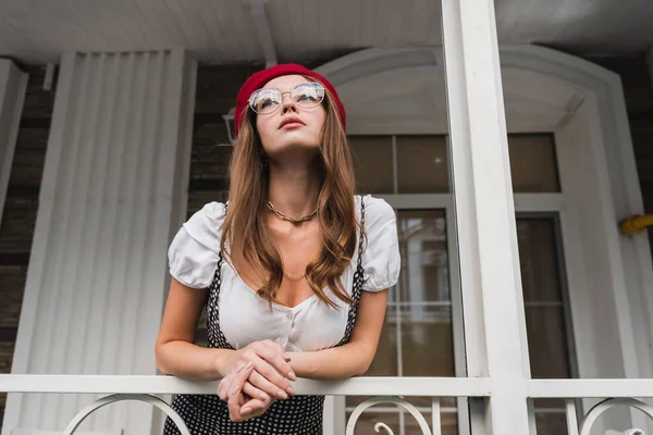 Jeune femme en béret rouge et lunettes debout sur le balcon et levant les yeux — Photo de stock