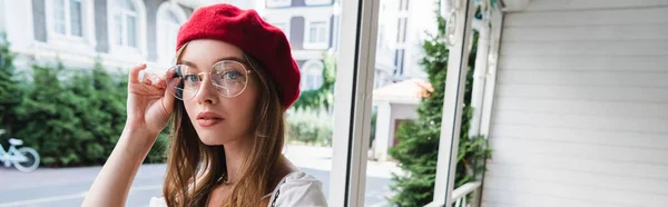 Jeune femme en béret rouge ajustant les lunettes et regardant la caméra, bannière — Photo de stock