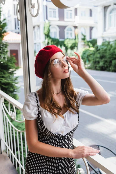 Jeune femme au béret rouge ajustant les lunettes et regardant loin sur le balcon — Photo de stock