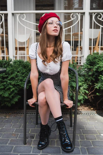 In voller Länge hübsche junge Frau mit roter Baskenmütze und Brille sitzt auf Stuhl draußen — Stockfoto