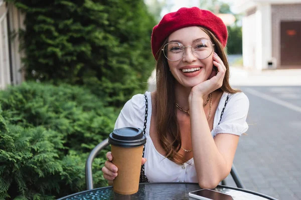 Joyeuse jeune femme en béret rouge et lunettes près de tasse en papier et smartphone avec écran blanc sur la table basse — Photo de stock