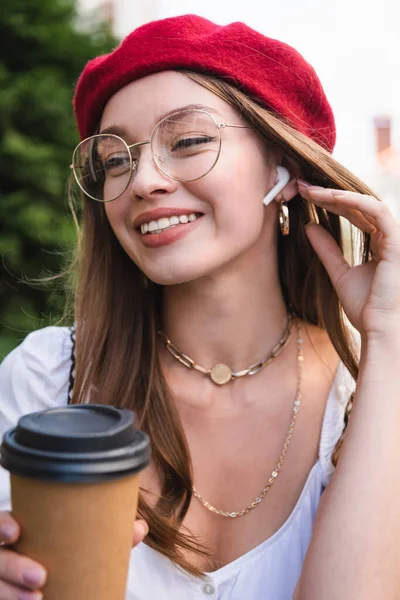 Heureuse jeune femme en béret rouge, écouteur et lunettes tenant tasse en papier — Photo de stock