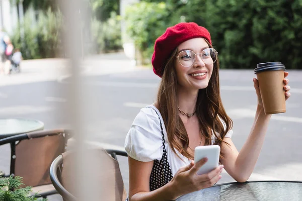 Mulher alegre em boina vermelha e óculos segurando copo de papel e smartphone no terraço do café — Fotografia de Stock