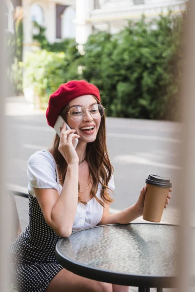 Cheerful woman in red beret and eyeglasses holding paper cup and talking on smartphone on terrace of cafe — Stock Photo