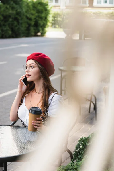 Young woman in red beret and eyeglasses holding paper cup and talking on smartphone on terrace of cafe — Stock Photo