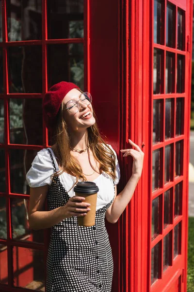 Радісна жінка в береті та окулярах тримає паперову чашку біля червоної телефонної кабіни — стокове фото