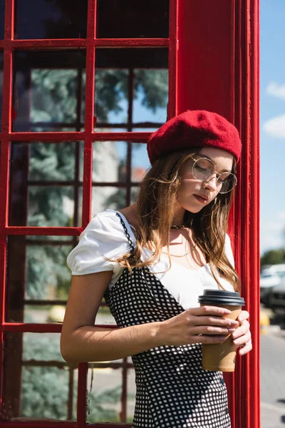 Jeune femme en béret et lunettes tenant tasse en papier près cabine téléphonique rouge — Photo de stock