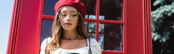 Donna gioiosa in berretto e occhiali vicino alla cabina telefonica rossa, striscione — Foto stock