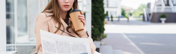 Vista ritagliata di giovane donna in trench in possesso di tazza di carta durante la lettura di giornale, banner — Foto stock