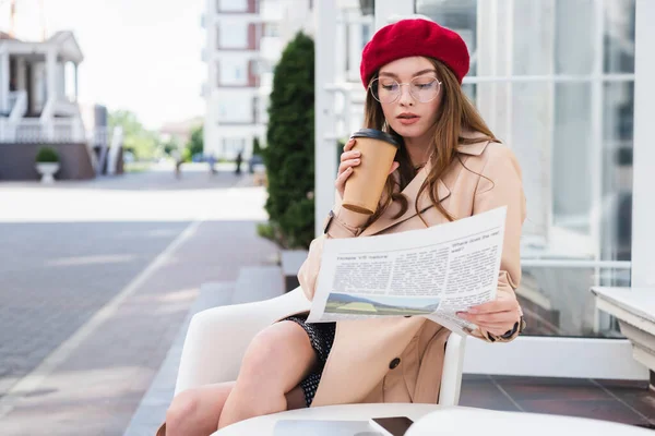 Молодая женщина в красном берете и бежевом плаще держа бумажную чашку во время чтения газеты — стоковое фото