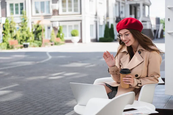Jovem feliz em boina vermelha e casaco de trincheira bege acenando mão durante chamada de vídeo no terraço do café — Fotografia de Stock