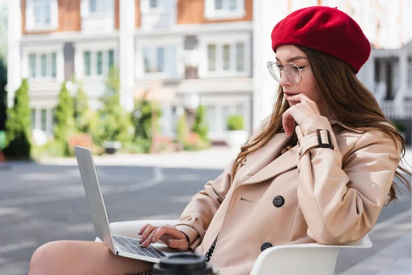 Joven freelancer en boina roja y gabardina beige usando laptop en terraza de cafetería - foto de stock