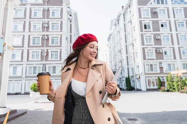 Радостная женщина в красном берете и бежевом плаще держа ноутбук и бумажную чашку улицы — стоковое фото