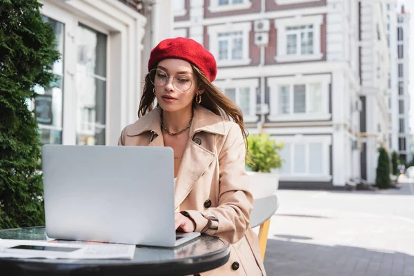 Pretty freelancer in eyeglasses, red beret and beige trench coat using laptop on terrace of cafe — Stock Photo