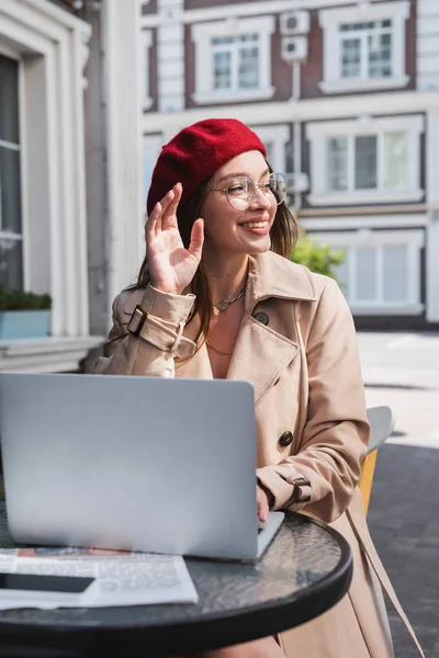 Feliz jovem mulher em boina vermelha acenando mão enquanto sentado no terraço do café perto de gadgets — Fotografia de Stock