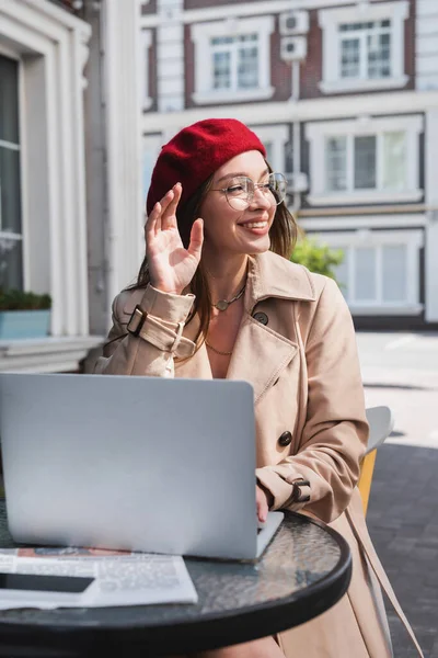 Felice giovane donna in rosso basco mano agitando mentre seduto in terrazza caffè vicino gadget — Foto stock