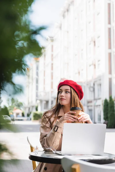 Pretty freelancer in red beret and beige trench coat holding paper cup near laptop on coffee table — Stock Photo