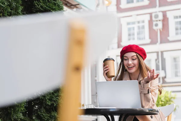 Happy freelancer in red beret holding paper cup and gesturing during video call in cafe — Stock Photo
