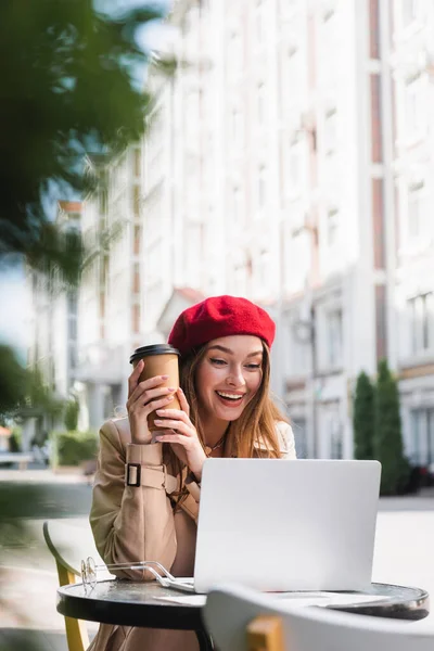 Cheerful freelancer in red beret and beige trench coat holding paper cup near laptop on coffee table — Stock Photo