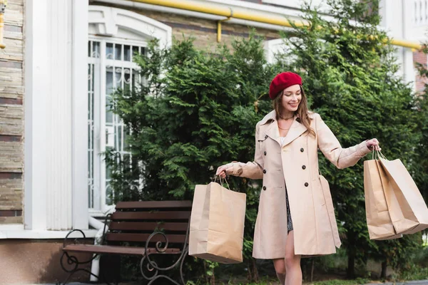 Joyeuse femme en béret rouge et trench coat beige tenant des sacs à provisions dans la rue urbaine — Photo de stock